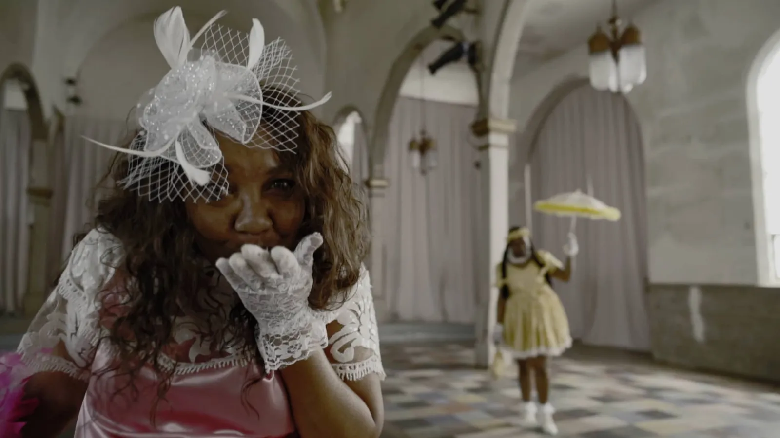 A black woman blows a kiss to the camera wearing a white lace dress with matching hat and gloves. Behind her is another Black woman in a yellow dress holding a matching parisol.