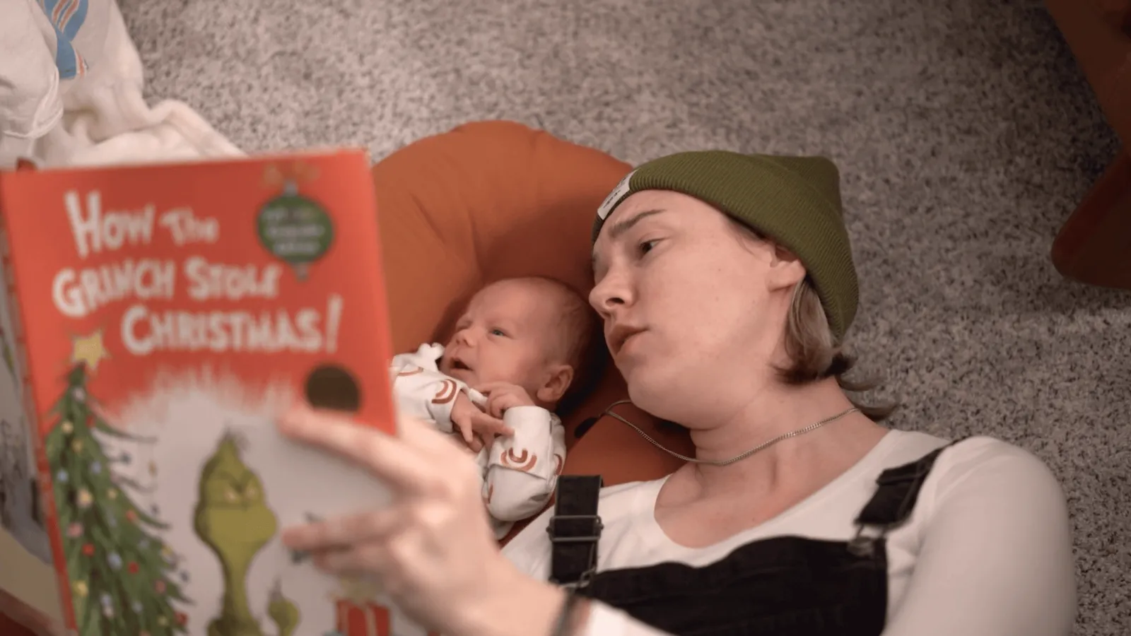 A white woman lays on the carpet reading a book to an infant.
