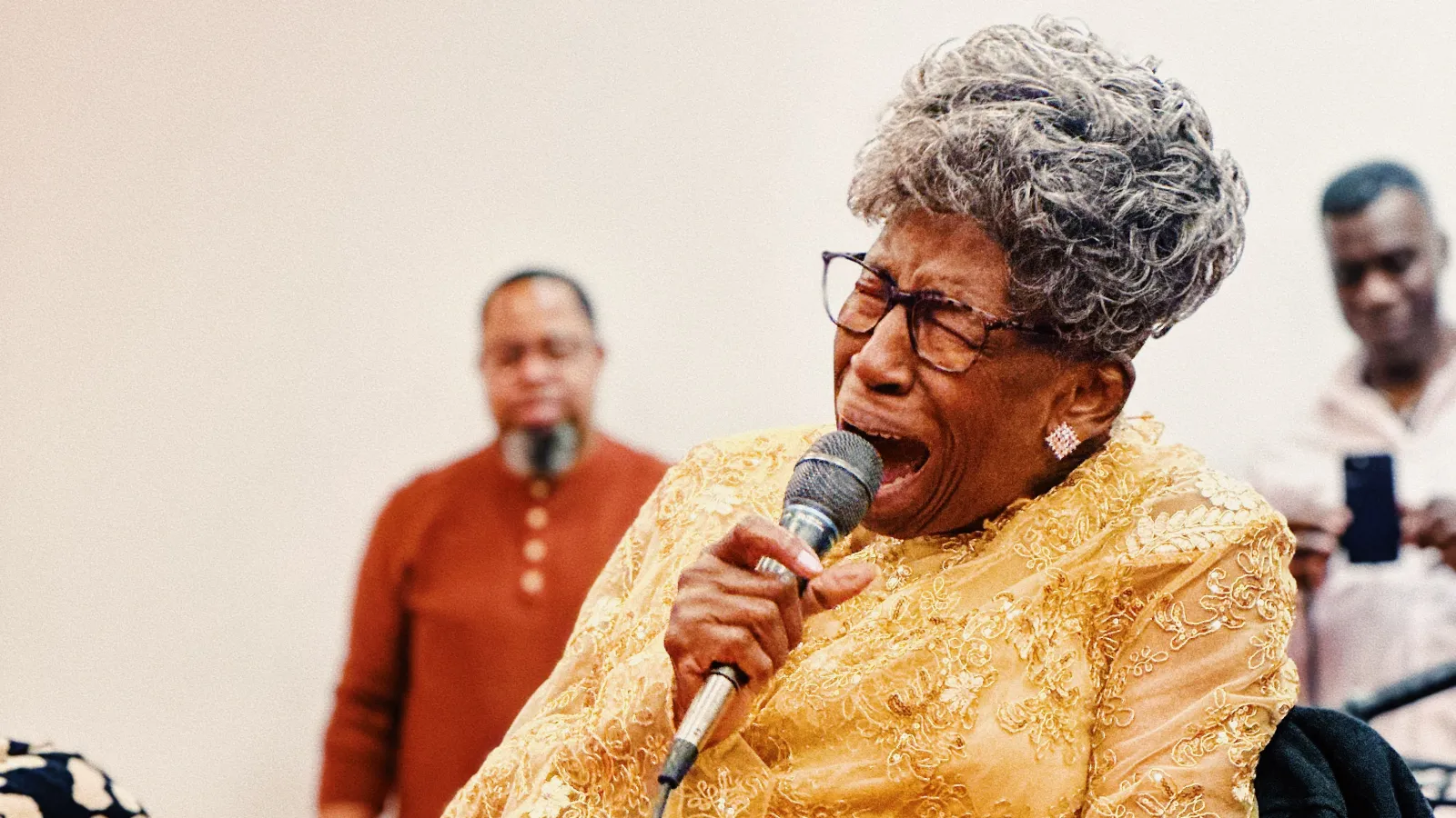 Mother Perry singing into a handheld microphone, wearing a gold long-sleeve dress.